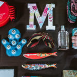 Um tira-senhas, dois computadores, uma luva de massagens, a letra M, uma máscara, duas sardinhas em porcelana, um perfume, uma caneca, uma mala e um copo com notas dentro