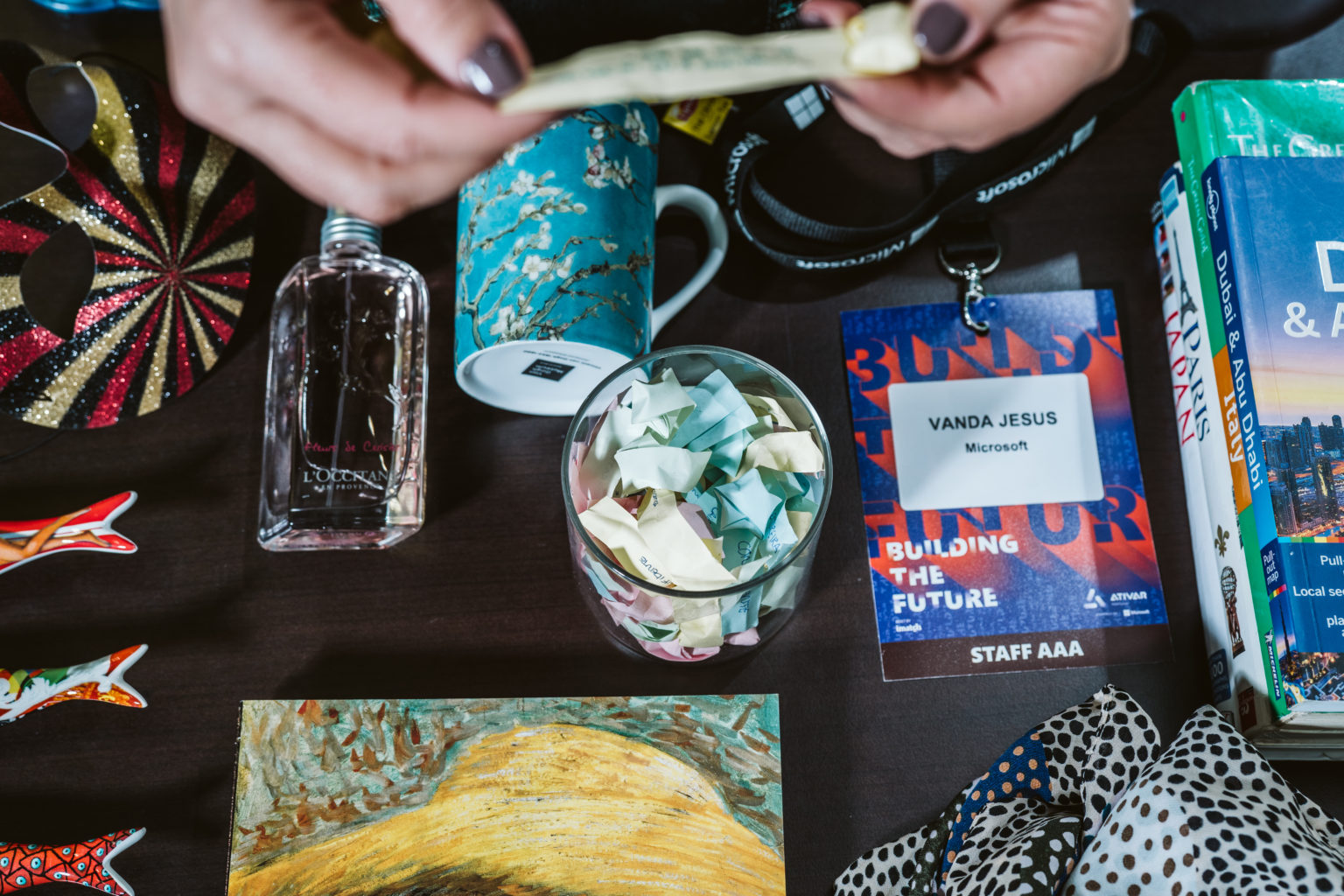 Umas mãos a agarrar um papel no ar e por baixo encontram-se uma máscara, um perfume, uma caneca, um copo comnotas dentro, um badge, uma coleção de livros de viagem, uma coleção de sardinhas em porcelana, um livro de pinturas e um lenço