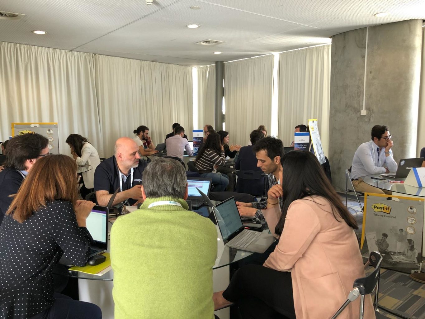 Vários grupos de pessoas sentados em mesas numa sala a participar no hackathon
