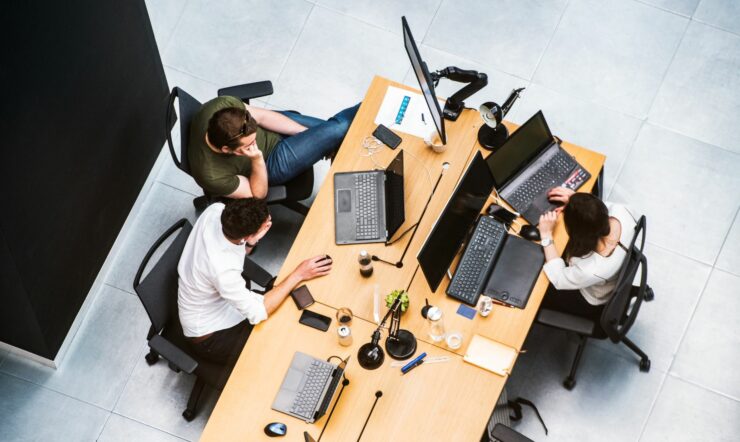 From above looking at three people sitting at a desk working on various laptops and screens
