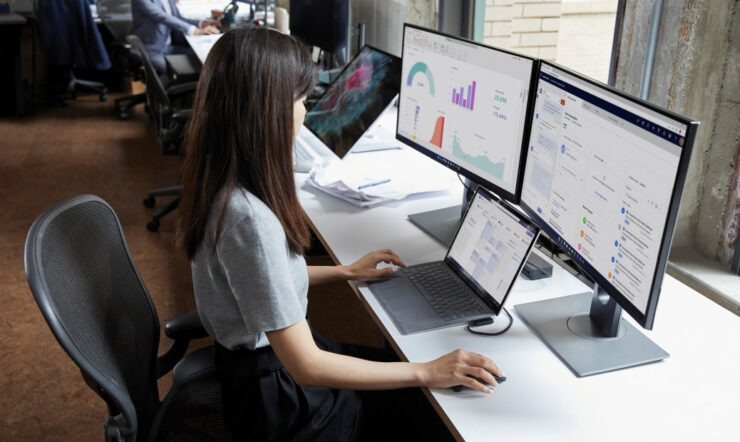 Woman sitting behind a desk looking at 2 screens and a surface device