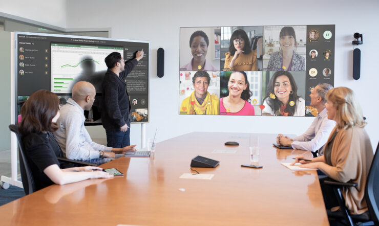Two woman and three men (one in a wheelchair) are hosting a coordinated meeting in a large conference room and are joined by several virtual attendees on a projection screen in Front of Room view.