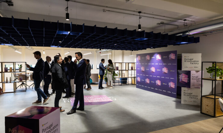 People inside of a room, speaking and walking around. The room is filled with tables, shelves and chairs.