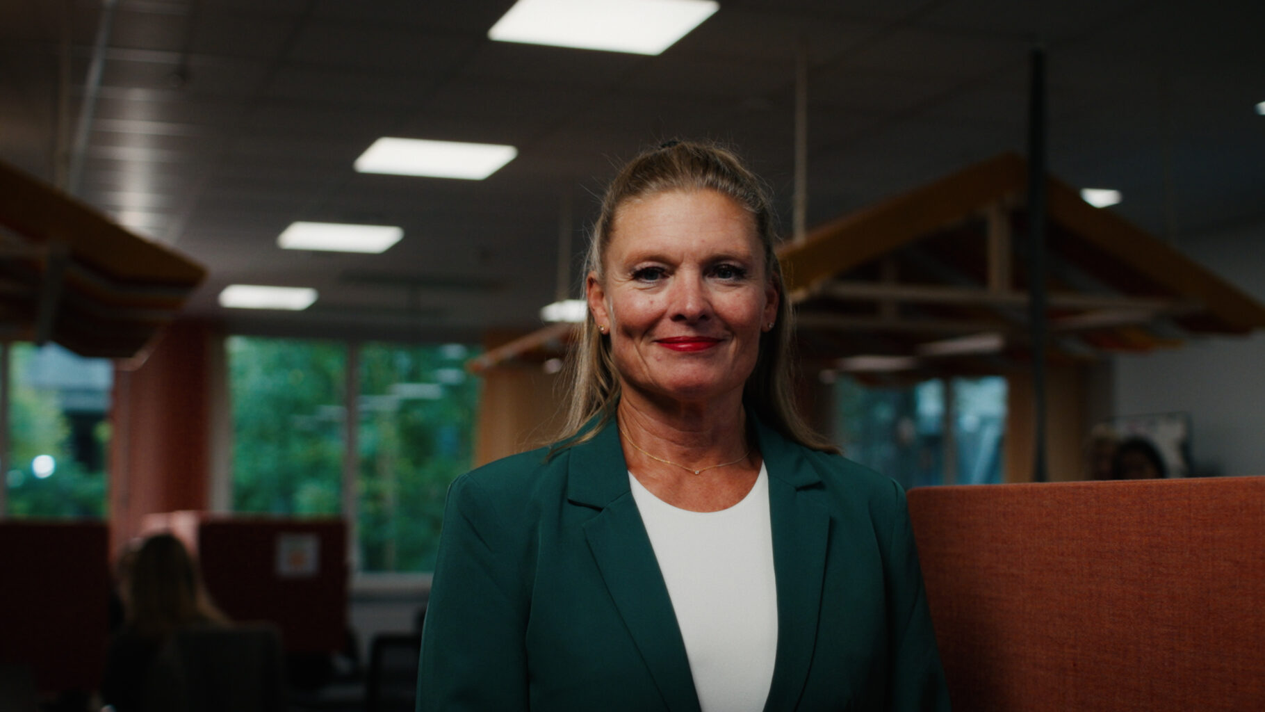 Person i grön kavaj står leende i ett kontor med varma färger.