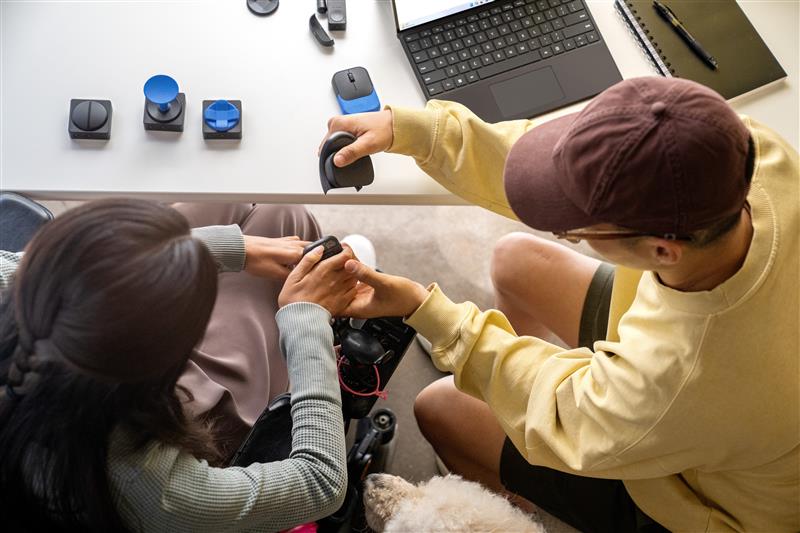 Kaksi henkilö työpöydän ääressä tutkimassa erilaisia saavutettavuusapuvälineitä