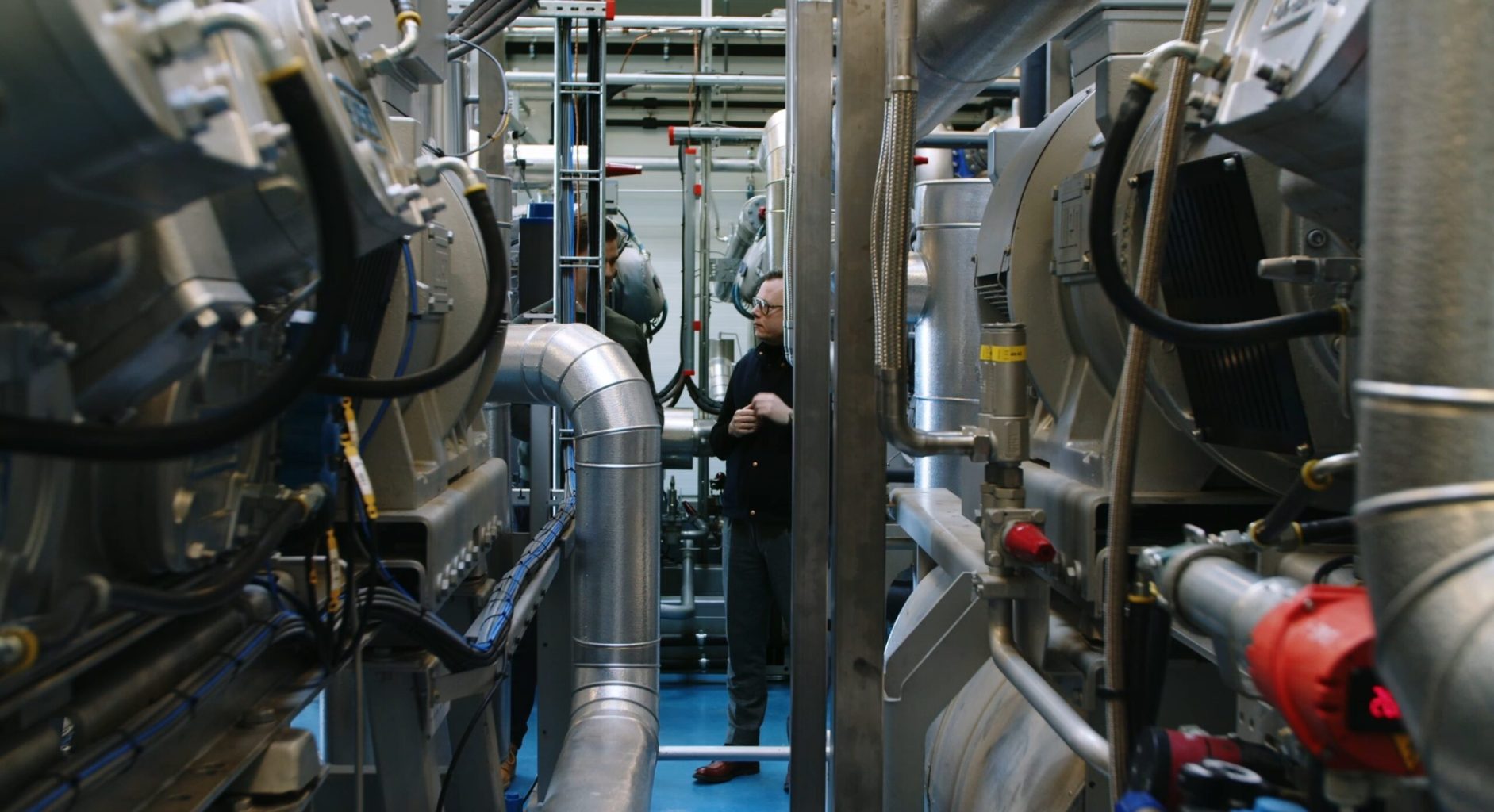 Inside an industrial facility with large pipes and machinery, a technician stands between equipment rows.