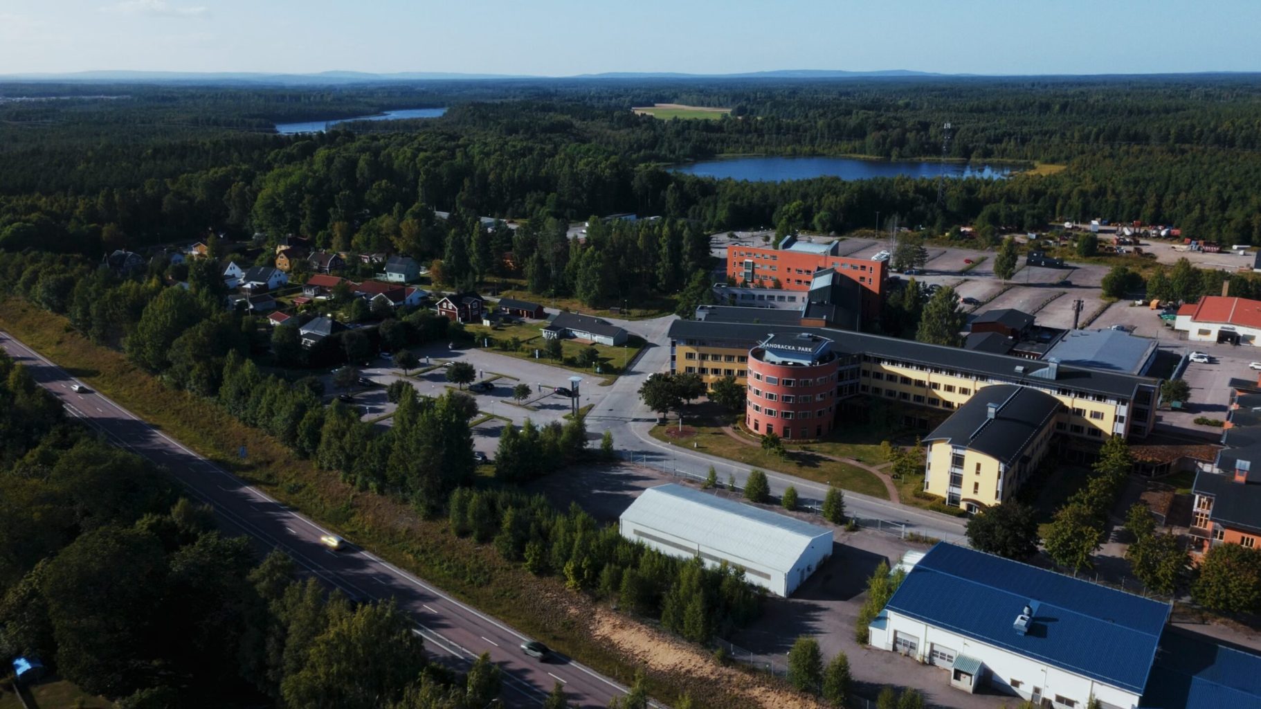 Flygbild över ett verksamhetsområde omgivet av skog och sjöar. Flera byggnader, parkeringsytor och en väg syns i förgrunden, med ett vidsträckt landskap i bakgrunden.