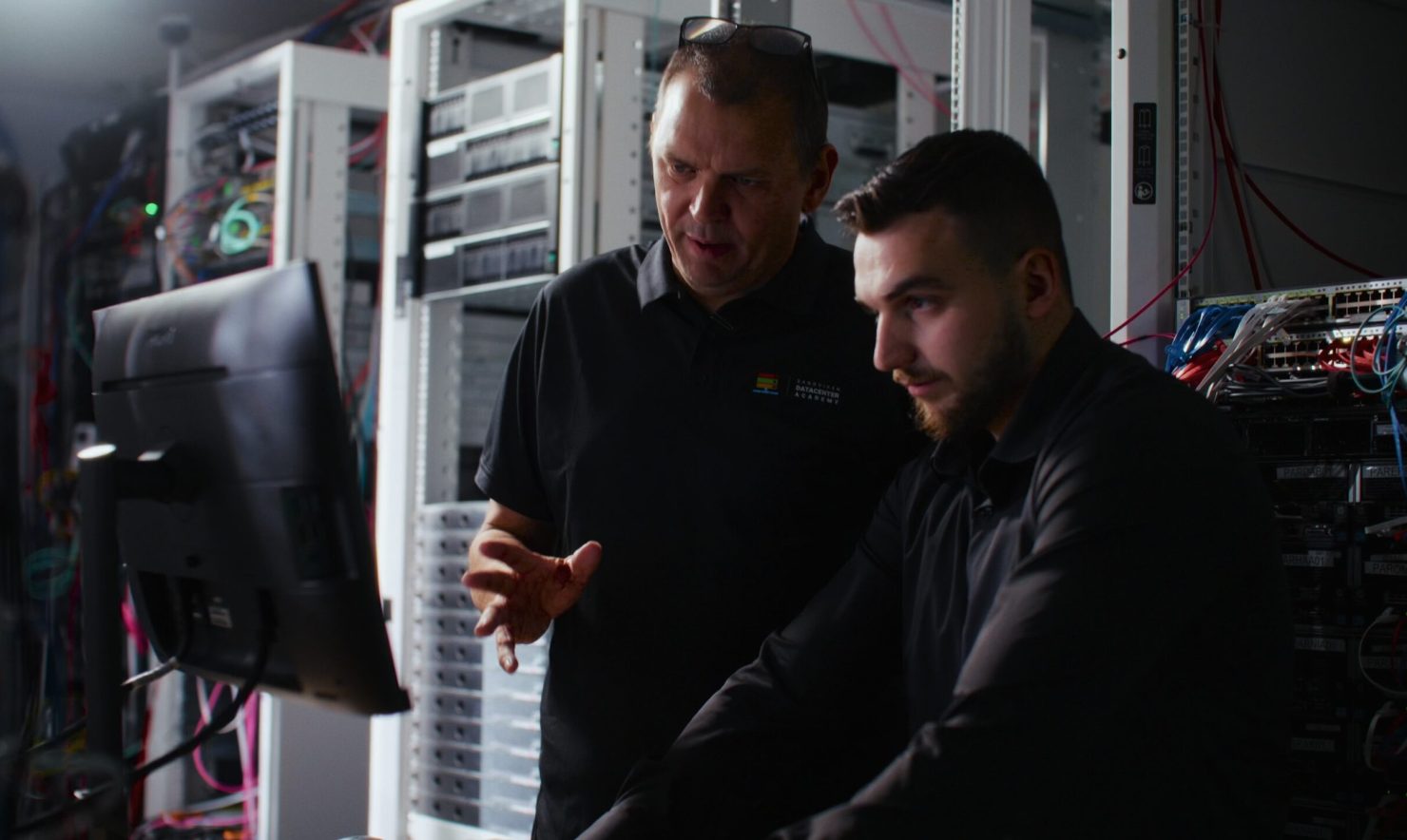 Två personer arbetar tillsammans framför en datorskärm i ett serverrum. Runt dem syns serverrack, nätverksutrustning och färgkodade kablar. Bilden är mörkt belyst med fokus på personerna och den tekniska miljön, vilket framhäver arbete med IT-infrastruktur och drift.
