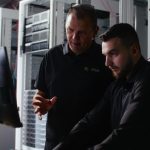 Två personer arbetar tillsammans framför en datorskärm i ett serverrum. Runt dem syns serverrack, nätverksutrustning och färgkodade kablar. Bilden är mörkt belyst med fokus på personerna och den tekniska miljön, vilket framhäver arbete med IT-infrastruktur och drift.